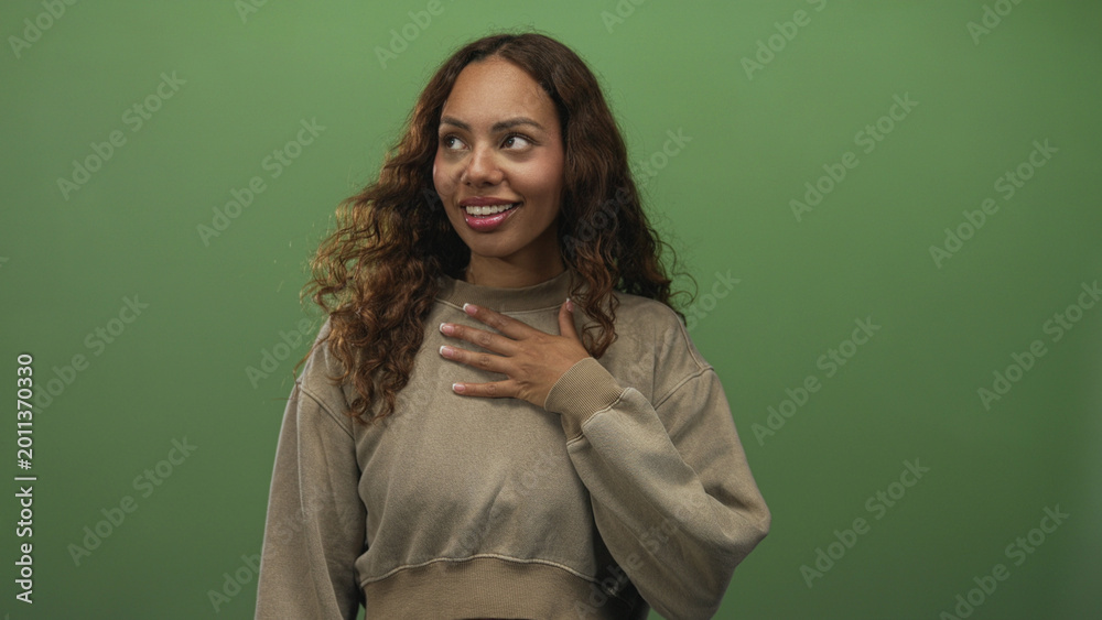 Obraz premium Young african american woman with curly hair and hand on chest smiling while wearing cropped sweater in green studio; surprise gratitude.