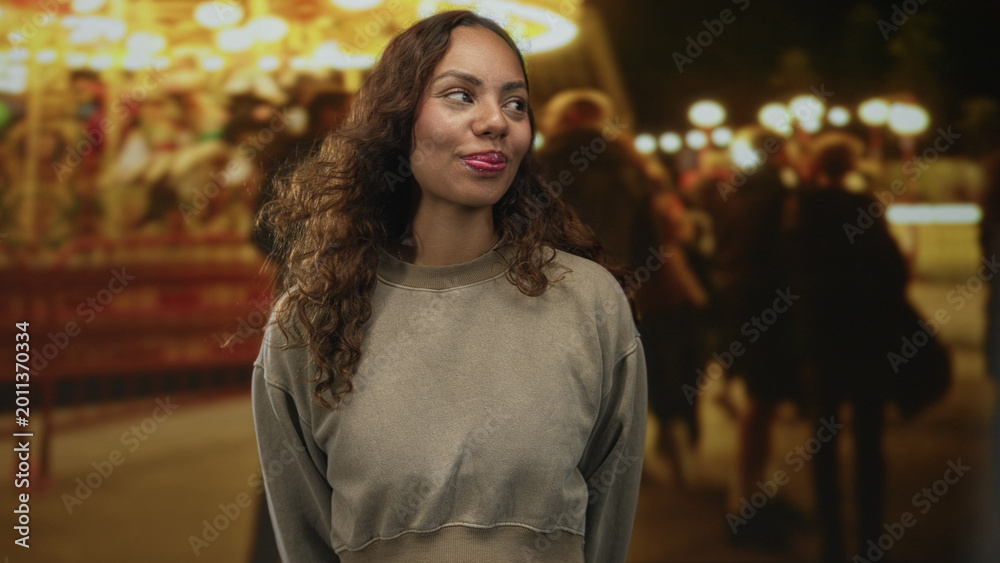 Fototapeta premium Woman smiling with closed eyes in a casual sweatshirt at an outdoor fair carousel, standing on a street at night; evening joy.