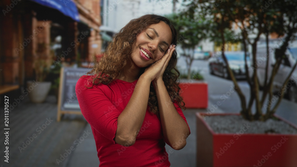 Fototapeta premium Young african american woman with hands together and cheek resting in sleeping gesture on busy city street; restful serenity.