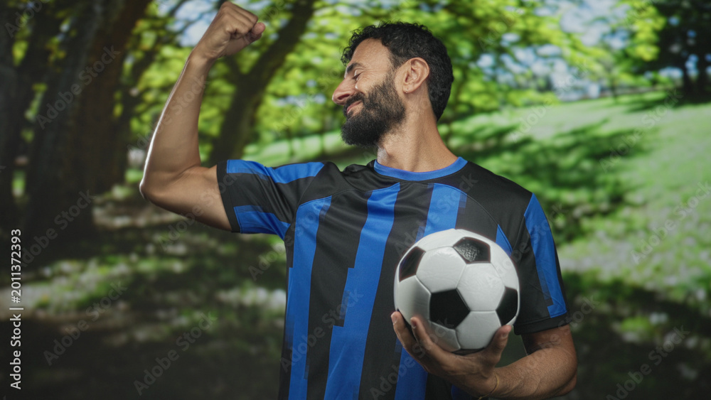 Obraz premium Man bearded soccer player in blue uniform holds soccer ball and flexes bicep while smiling in forest; pride.