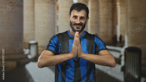 Fotografi Man in blue and black soccer jersey with hands pressed together praying in building corridor; penitence reflection