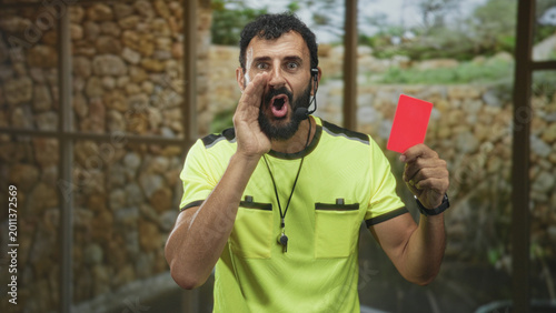 Valokuva Referee man holding a red card, hand cupped at mouth and shouting into a headset inside a building; authority