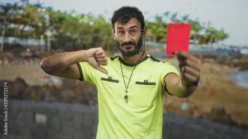 Fotografie Referee man in yellow shirt shows red card and thumbs down gesture with whistle and headset visible on street; discipline authority