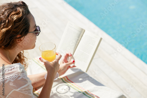 Billede på lærred Woman reading book and drinking juice by poolside in summer