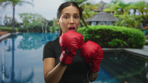 Bild auf Leinwand Hispanic woman boxer with red gloves in guard with open mouth at resort pool building; determination training