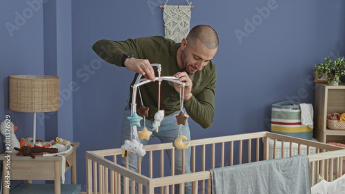 Foto Man adjusts crib mobile with his hand while bending over a wooden cradle and checking hanging toys in a studio baby room; tenderness