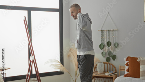 Bild auf Leinwand Man pulls hood up on hoodie while glancing at mirror by the bed in a studio bedroom with pampas grass and rattan chair; quiet contemplation