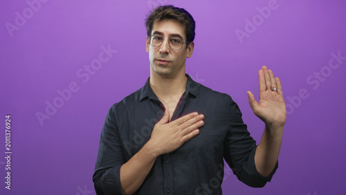 Bilde på lerret Man wearing glasses and dark shirt, hand placed over chest and other hand raised in oath gesture in studio purple backdrop with wedding ring visible; sincerity