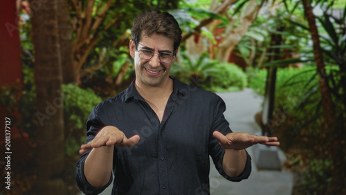 Canvastavla Man using outstretched hands to gesture while smiling on a planted walkway beside a building, wearing glasses and dark shirt with visible palms; amusement