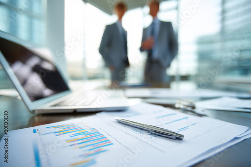 Close-up of private equity partners reviewing financial reports on a modern office desk, two professionals discussing investment charts on a laptop and printed documents, confident business atmosphere