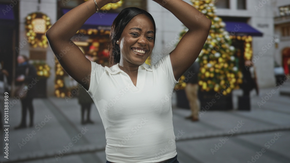 Obraz premium Black woman raising arms above head in white polo smiling on a busy city street with holiday lights and a decorated storefront; holiday joy.