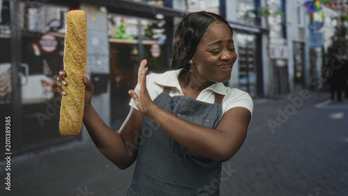Photos Woman baker in apron holds a baguette in her left hand and recoils with her right hand in a disgusted grimace on a street; disgust hesitation
