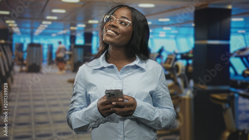 Konstfotografi Woman holding smartphone with both hands, smiling and glancing up in a gym filled with treadmills and machines; contentment focus