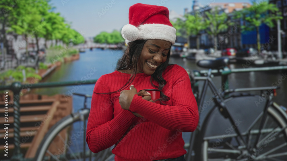 Naklejka premium Woman in santa hat smiles and clasps hands by a canal bridge with bicycle on a street; holiday joy warmth.