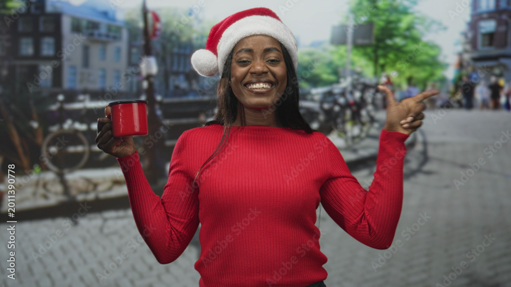 Naklejka premium Woman in santa hat holding a red mug and pointing finger on a cobbled street with bicycles and buildings, wearing red sweater and smiling at camera; holiday joy.