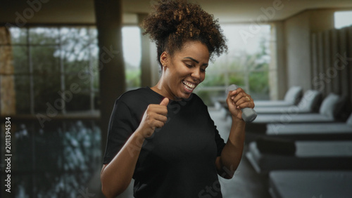 Foto Woman young african american smiling and holding dumbbell in spa building lounge with poolside beds and large windows; joyful fitness