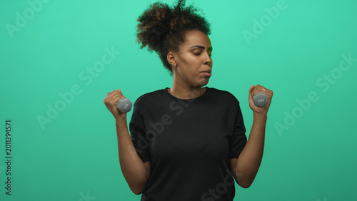 фотографія Woman holding small dumbbells and lifting forearms in green studio while wearing black shirt and curly hair pulled up; fitness effort