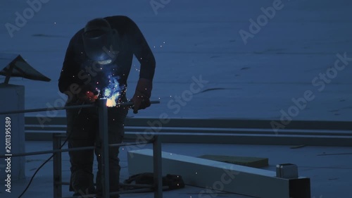 Professional welder in protective gear carefully joins metal components on flat roof surface at dusk, orange sparks illuminate work area highlighting industrial activity. Industrial process, hard work