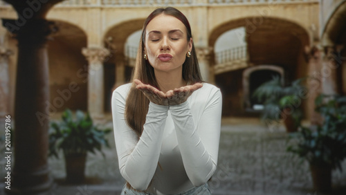 Foto Woman blowing a kiss with cupped hands showing palms and fingers, wearing white top in a historic courtyard building; welcome greeting joy