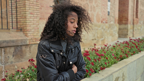 Konstfotografi Teen woman with curly hair stands with arms crossed beside a stone building and red flowerbed; melancholy solitude