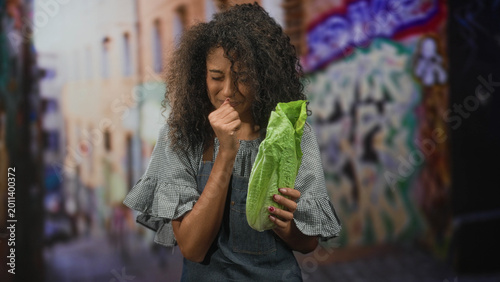 Photos Woman with curly hair wearing apron points finger to nose while holding romaine lettuce in graffiti lined street, grimacing at smell; disgust food caution