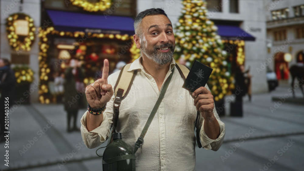 Fototapeta premium Man pointing finger while holding passport on a decorated street with christmas tree and lights; holiday travel joy.