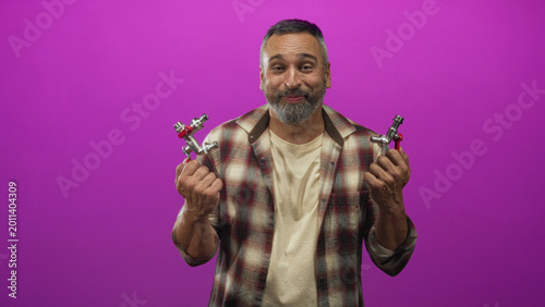 Bild auf Leinwand Man holding two faucet valves with both hands in a magenta studio, wearing a flannel shirt with grey beard and short grey hair; playful repair