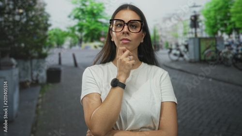 Schilderij op canvas Woman with long brunette hair wearing white t shirt and oversized glasses, smartwatch visible, hand on chin on a cobblestone street with bicycles nearby; contemplation