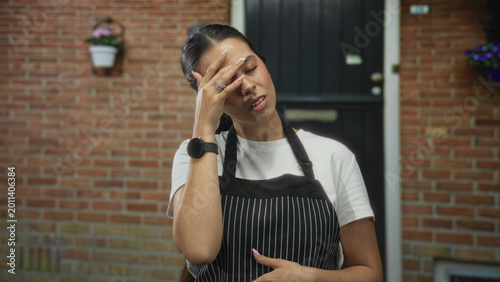 Bild auf Leinwand Young woman in striped apron touches throat and presses forehead with hand at building entrance by a black door and brick wall; discomfort worry