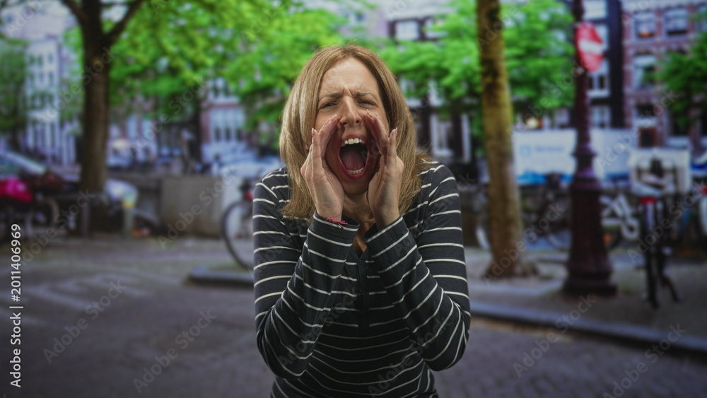 Fototapeta premium Woman cupping hands and shouting with open mouth on an urban street near bicycles and trees, wearing striped long sleeve top; alarm protest anger.