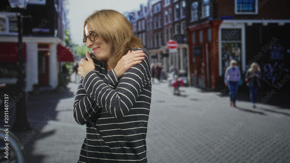 Fototapeta premium Woman hugs herself with crossed arms wearing glasses and striped sweater on a sunlit street; self comfort serenity.