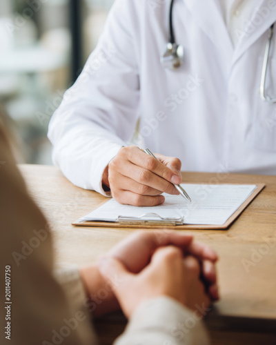 A doctor wea a white coat and stethoscope is writing on a clipboard while consulting with a patient sitting across from him at a desk.