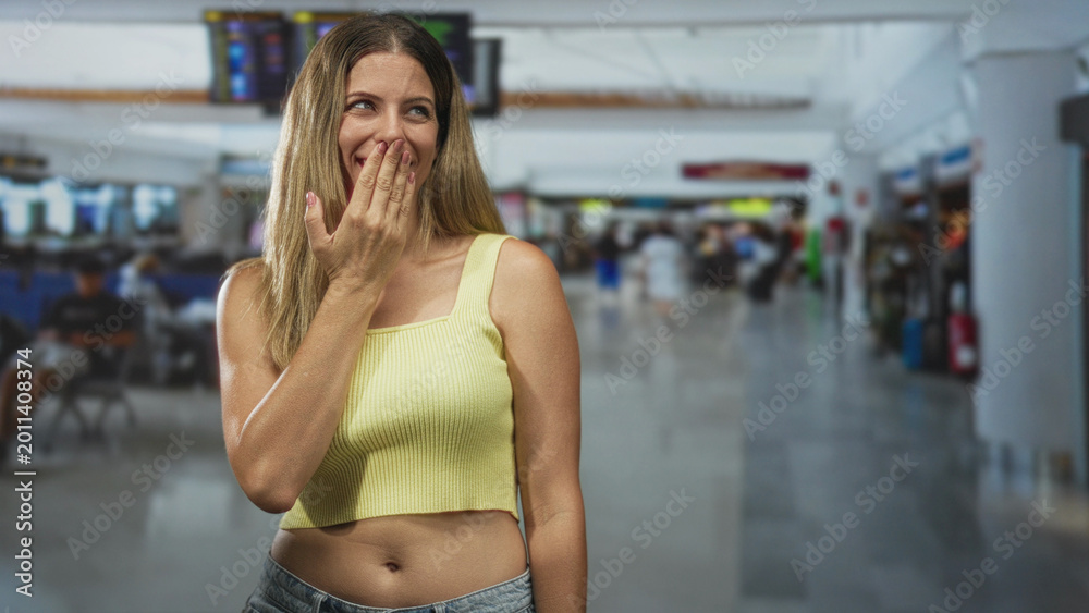 Fototapeta premium Woman covering mouth with hand in airport terminal wearing yellow crop top and jeans showing midriff; playful warmth.