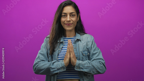 Schilderij op canvas Woman young with hands pressed together in prayer, wearing denim jacket and striped crop top in a studio with purple backdrop; serenity