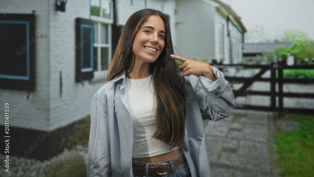 Fototapeta premium Young hispanic woman points fingers to cheeks and smiles in front of building by a wooden gate and stone path; joy confidence.