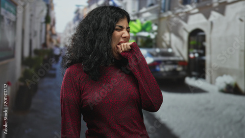 Tableau sur toile Young brunette woman in red sweater bites her fist on a narrow city street, hand partly in mouth while suppressing a yawn and closing eyes; tired solitude