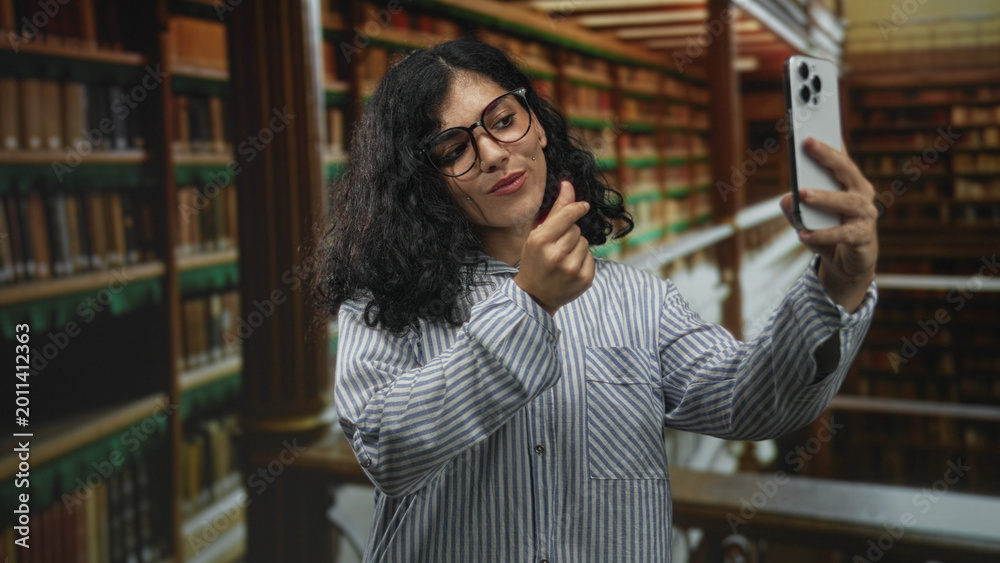 Fototapeta premium Young woman holding smartphone and making finger heart gesture in building library; self expression confident.
