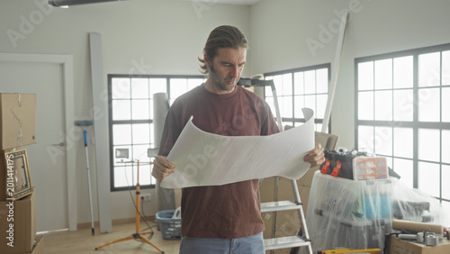 Photos Man holding large blueprint, furrowed brow, inspecting plans in cluttered room with boxes inside building; uncertainty home planning