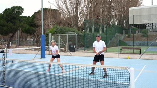 Active mature couple engaged in a dynamic game of pickleball on a blue outdoor court. The woman hits the ball as her partner anticipates the next move