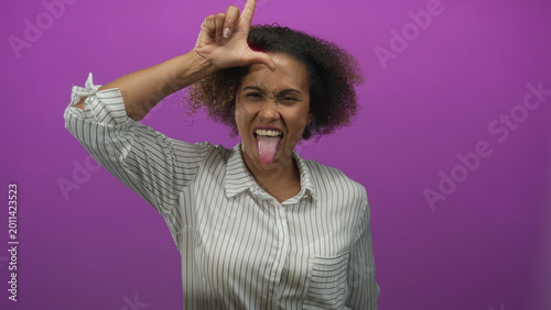 Принти на полотні Woman sticking out tongue and making loser l sign with hand on forehead in a purple studio; playful mischief