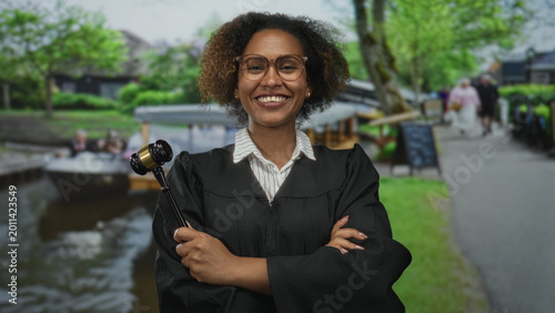 Cuadro en lienzo Woman judge smiling and holding gavel with arms crossed on street by canal in amsterdam; confidence authority law