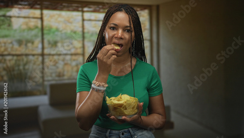Tableau sur toile Woman holding glass bowl of chips with finger to lips for silence in a building living room; thoughtful pause