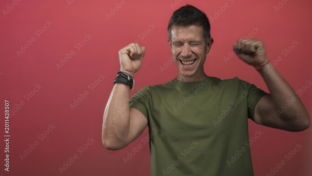 Fototapeta premium Man smiling and raising both fists, showing forearms and bracelets against a vivid red studio wall; joy celebration.