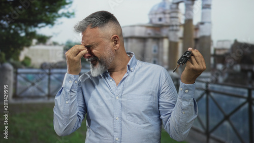 Φωτογραφία Man pinches nose while holding car keys on an old street with stone buildings and metal railing; disgust misplacement