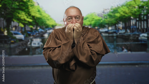 Картината върху платно Man monk covers mouth with hands in street by a canal, brown robe visible and eyes closed in a prayerful posture; contemplation devotion