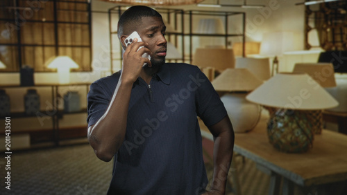 Photography Man holding phone to ear with tense, attentive expression among table lamps, lampshades and decorative vases in a furniture showroom; concern urgent call