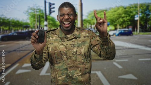 Photos Soldier man in camouflage uniform smiles, holds a walkie talkie and flashes rock horns while standing at a busy street intersection; pride duty