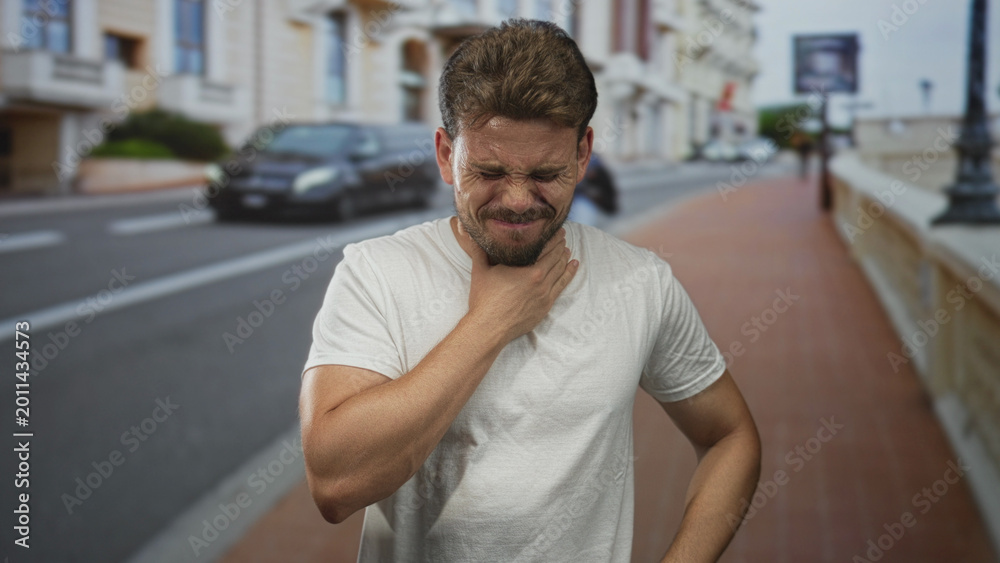 Fototapeta premium Young hispanic man clutching his neck and grimacing while standing on a city street wearing a white t shirt and squinting in daylight; sore throat pain.