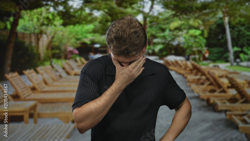 Canvastavla Man presses hands to face in outdoor poolside area with wooden lounge chairs and trees on a street walkway; distress