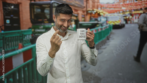 Fotografía Man holding a mug that reads i am the boss while pointing index finger in busy street with green barriers and parked van; confidence humor